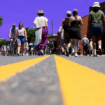 People walk and bike down the middle of the road during Boston's Open Streets event. The sky is tinted purple.