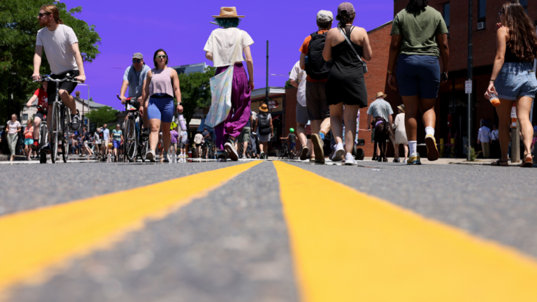 People walk and bike down the middle of the road during Boston's Open Streets event. The sky is tinted purple.