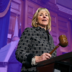 Massachusetts Senator Karen Spilka stands at a podium in the senate chambers holding a gavel. The background is tinted purple.