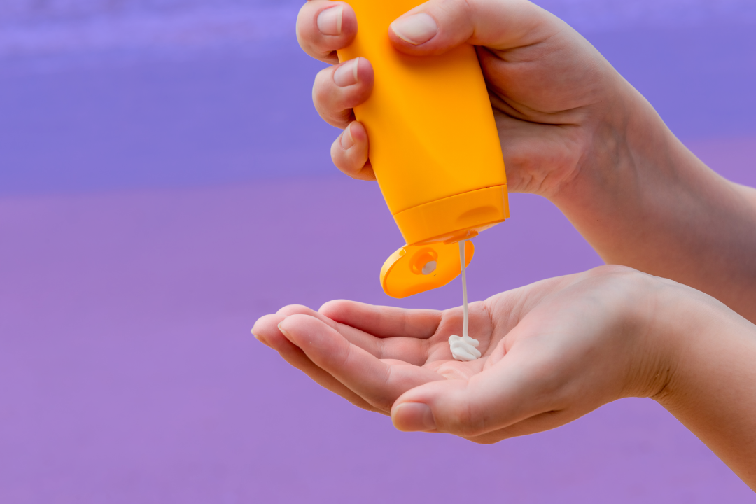 A hand squeezes sunscreen from an orange bottle onto another open hand. The background is a beach scene tinted purple.
