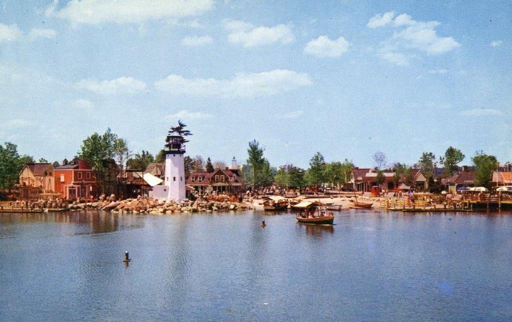In a vintage photo from the mid-20th century, a body of water is shown with a small boat floating in the water. Several buildings can be seen on the shore, in addition to a white lighthouse.