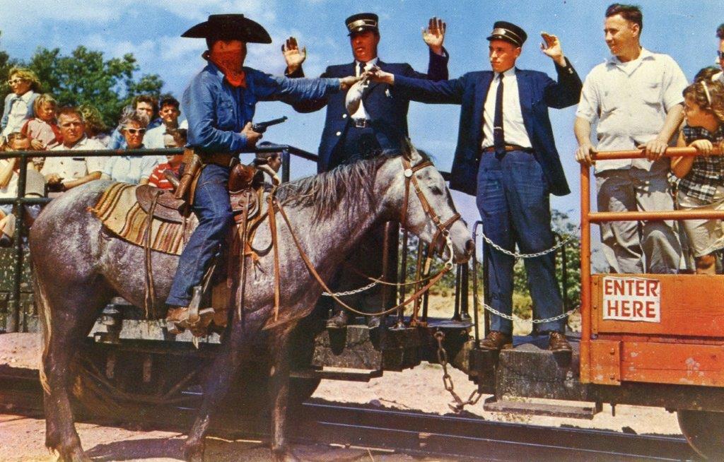 In a vintage photo from the mid-20th century, a man on a horse is shown wearing Old West themed clothing and a bandana around his face. He is pointing a gun at two men in train conductor costumes, who are shown handing him a pouch.