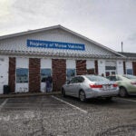 Three parked cars and two people stand outside a Massachusetts Registry of Motor Vehicles building