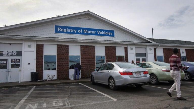 Three parked cars and two people stand outside a Massachusetts Registry of Motor Vehicles building