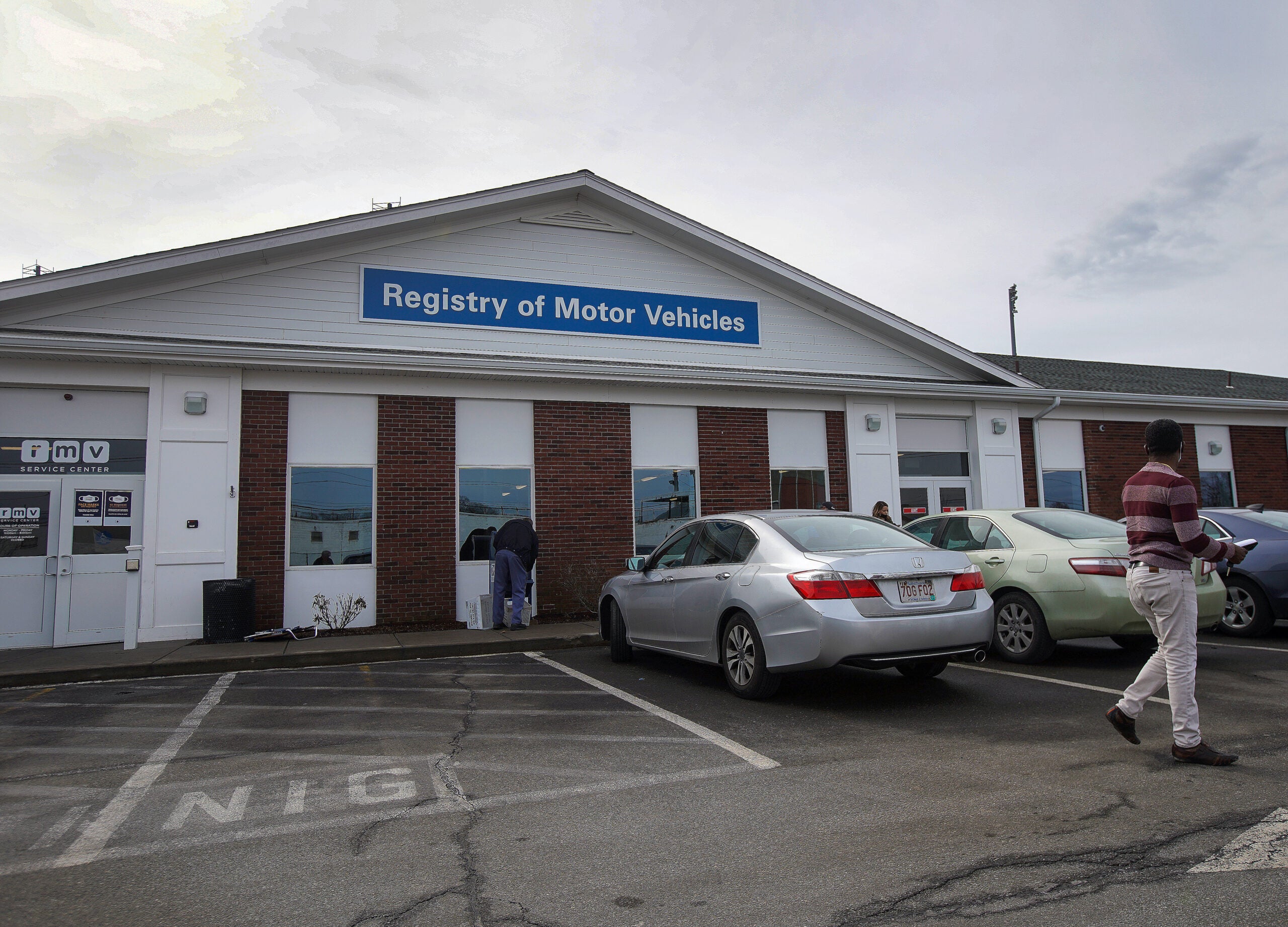 Three parked cars and two people stand outside a Massachusetts Registry of Motor Vehicles building