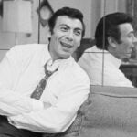 Ed Ames in a white button up shirt and tie, smiling and sitting on a couch with his arms folded as he leans back against a mirror.