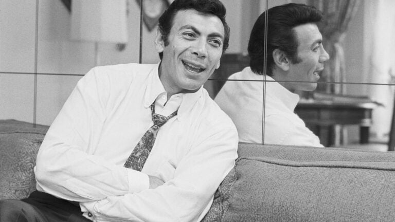 Ed Ames in a white button up shirt and tie, smiling and sitting on a couch with his arms folded as he leans back against a mirror.