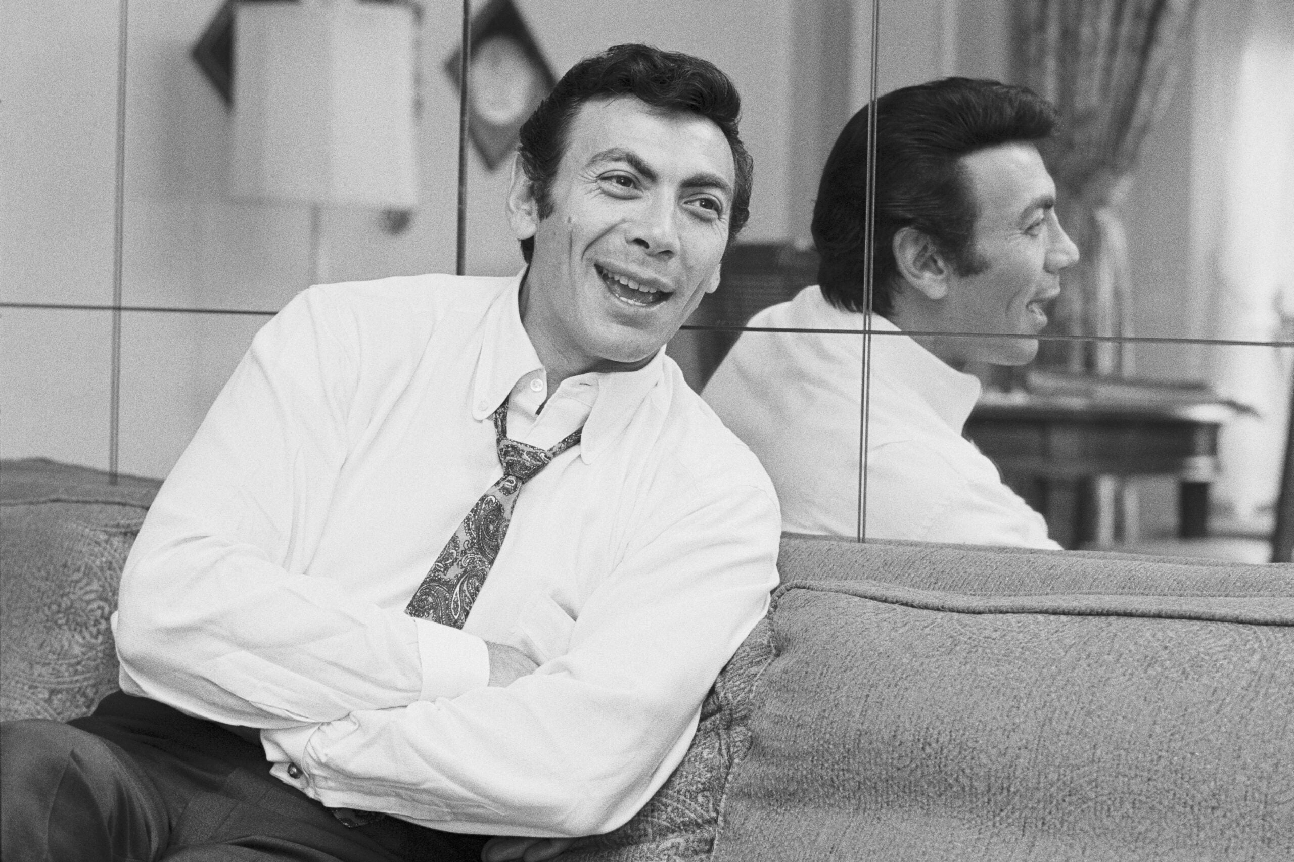 Ed Ames in a white button up shirt and tie, smiling and sitting on a couch with his arms folded as he leans back against a mirror.