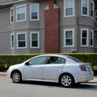 A photo taken near the scene of a fatal Waltham shooting Monday. A silver car is shown parked on the street in front of a house, with its lefthand rear window shattered.
