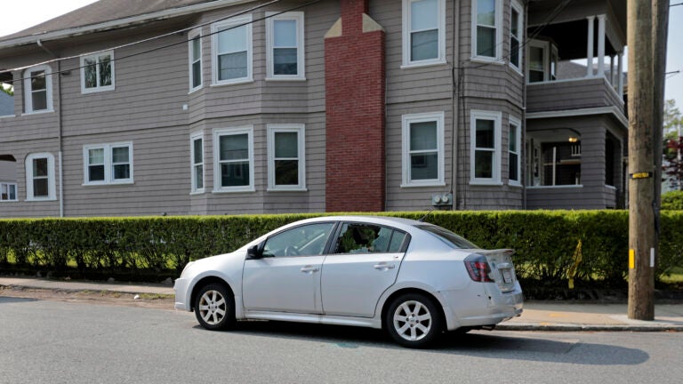 A photo taken near the scene of a fatal Waltham shooting Monday. A silver car is shown parked on the street in front of a house, with its lefthand rear window shattered.