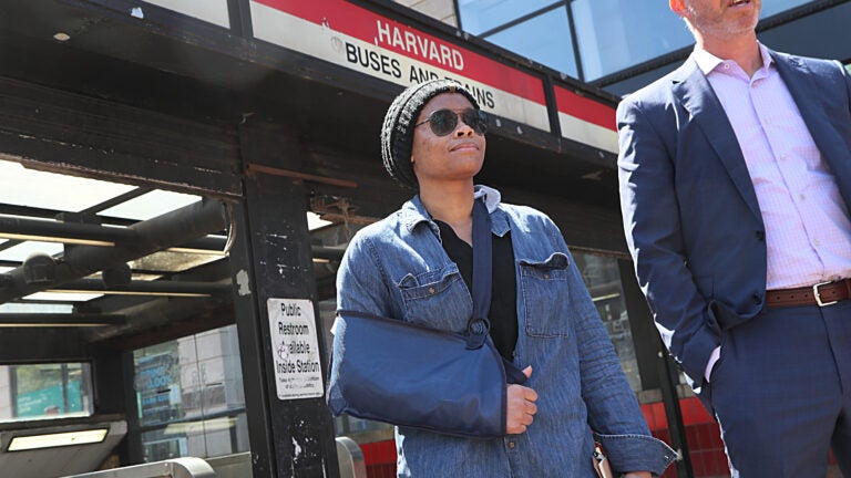 Joycelyn Johnson is pictured in front of Harvard Station. She is wearing a blue denim jacket, a knit beanie, and dark sunglasses. Her right arm is in a sling.