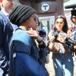 Joycelyn Johnson stands in the middle of a small crowd of people outside a T station. She is wearing a denim jacket, sunglasses, and a knit beanie, and her right arm is in a sling. She is gesturing to her right shoulder.