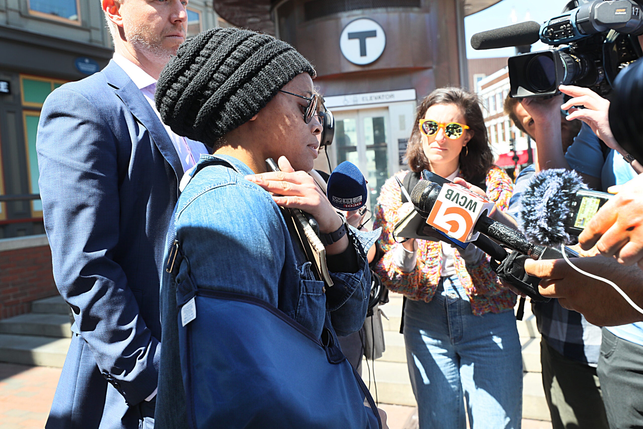 Joycelyn Johnson stands in the middle of a small crowd of people outside a T station. She is wearing a denim jacket, sunglasses, and a knit beanie, and her right arm is in a sling. She is gesturing to her right shoulder.