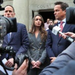 Karen Read, center, in a gray suit, stands on the steps of the courthouse, flanked by two of her lawyers. They are surrounded by members of the media holding cameras and microphones.