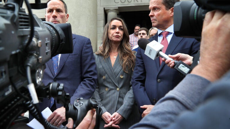Karen Read, center, in a gray suit, stands on the steps of the courthouse, flanked by two of her lawyers. They are surrounded by members of the media holding cameras and microphones.