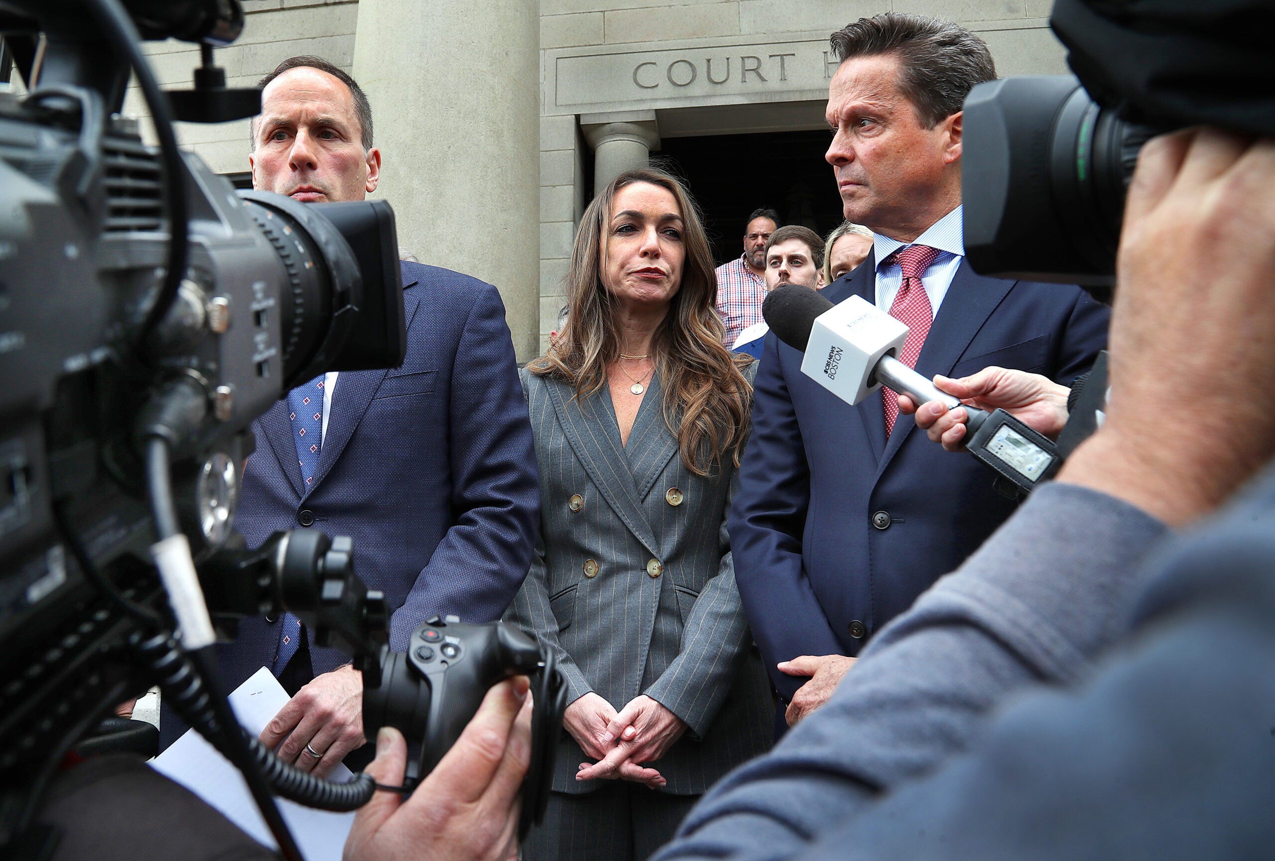 Karen Read, center, in a gray suit, stands on the steps of the courthouse, flanked by two of her lawyers. They are surrounded by members of the media holding cameras and microphones.
