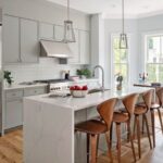 A kitchen with smooth-front gray cabinetry that goes to the ceiling, an island with waterfall edges and seating for four, hardwood flooring, two pendant lights, and a windowed breakfast nook. This photo is used to illustrate open houses going on this weekend.