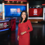 Jennifer Peñate stands in a TV news studio in front of monitors bearing WCVB's logo. She is wearing a red long-sleeved dress and smiling at the camera. Her black hair is worn loose.