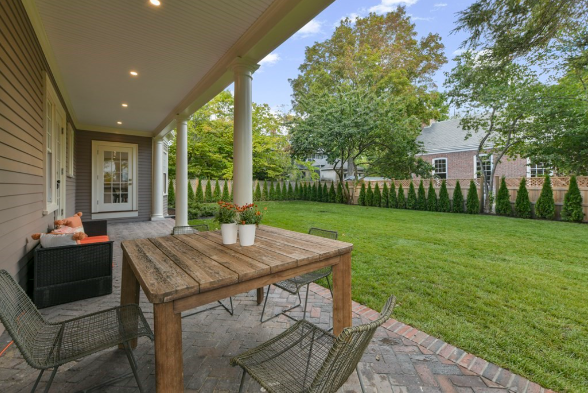 A brick patio sits under the covered porch, looking into the yard lined with trees.