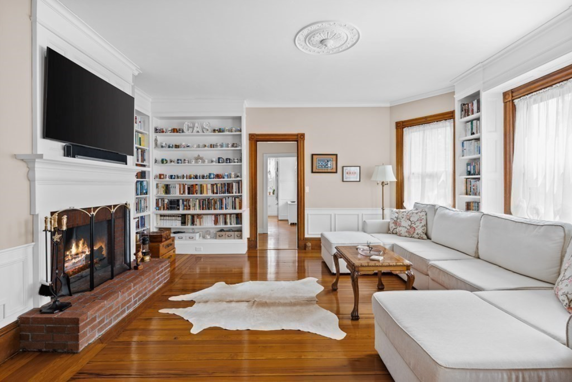 Living room with built-in bookshelves, bay window, gas fireplace set in brick, hardwood floors, and beige walls.