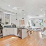 Living room and kitchen connected via open floor plan. The space has pendant lighting, hardwood floors, and single-hung windows.