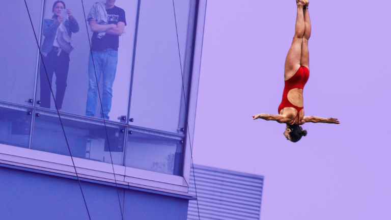 A woman in a red bathingsuit is upside down diving through the air into the Boston Harbor. People from a nearby building look on. The background is tinted purple.