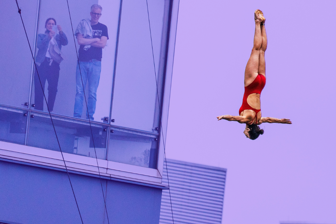 A woman in a red bathingsuit is upside down diving through the air into the Boston Harbor. People from a nearby building look on. The background is tinted purple.