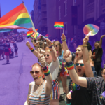 People dressed in rainbow clothing wave rainbow Pride flags along a Pride parade route in Boston. The background of the image is tinted purple.