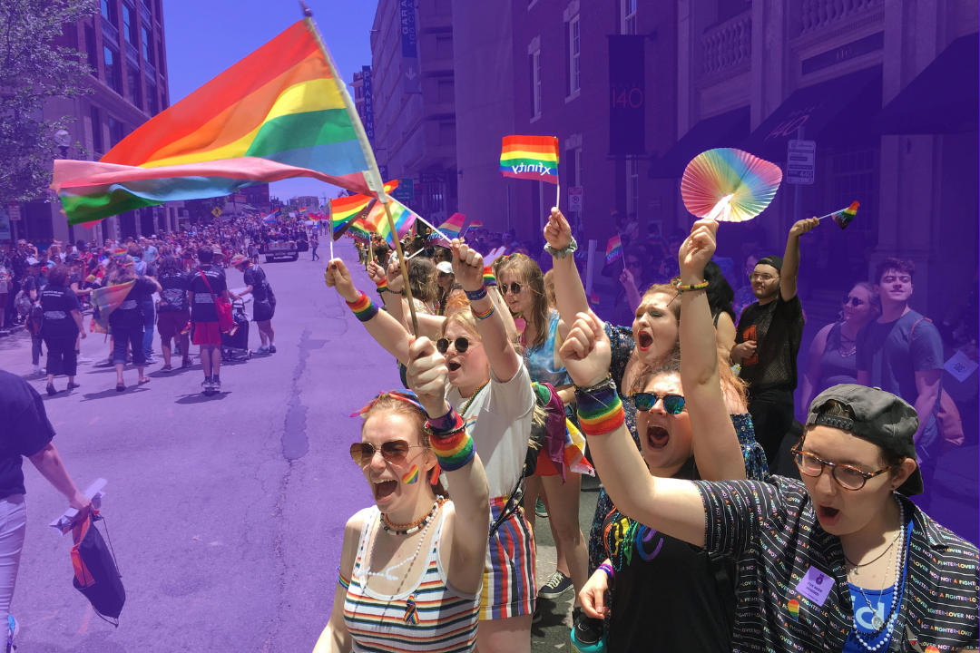 People dressed in rainbow clothing wave rainbow Pride flags along a Pride parade route in Boston. The background of the image is tinted purple.