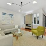 a room with modern furnishings: light-gray walls, a gray sectional, a chartreuse winged armchair, a horizontal gas fireplace, wood flooring, and long horizontal windows. The light fixture over it all looks like streetlights extended on three long arms.