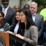 Boston Mayor Michelle Wu stands at a lectern during a press conference. She is surrounded by people, wearing her hair down, smiling, wearing a gray cardigan.