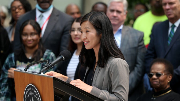 Boston Mayor Michelle Wu stands at a lectern during a press conference. She is surrounded by people, wearing her hair down, smiling, wearing a gray cardigan.
