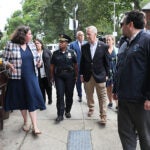 Several Boston politicians and officials tour a South Boston public housing complex outdoors on foot. They are pictured in various states of business or work attire, mid-conversation.