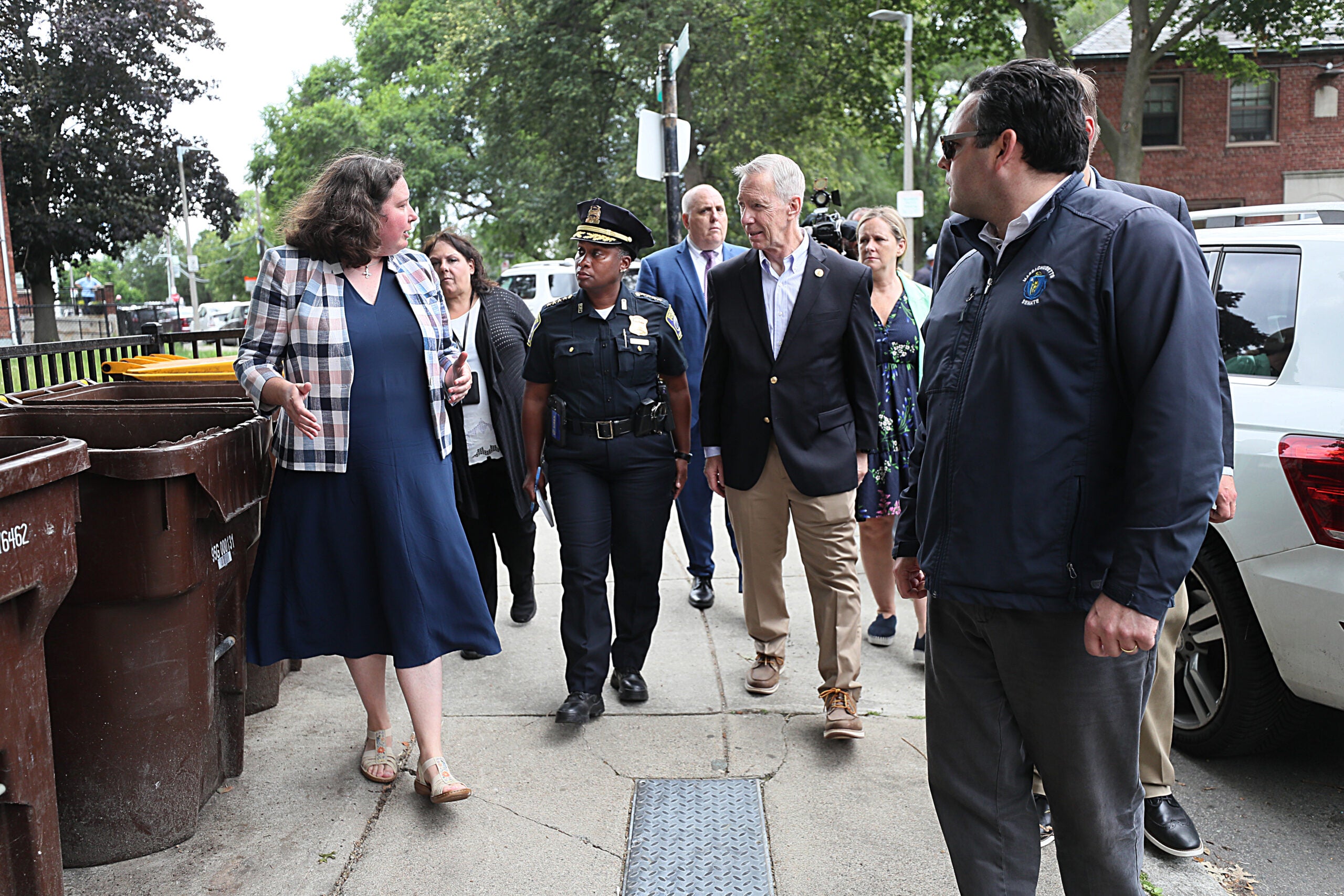 Several Boston politicians and officials tour a South Boston public housing complex outdoors on foot. They are pictured in various states of business or work attire, mid-conversation.