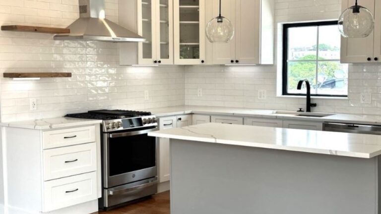 A kitchen with shiny, white subway tile, a gray island with a marble-like countertop, two glass globe pendant lights, stainless steel appliances, open shelving, white Shaker-style cabinets (a few with glass doors), a window with a black frame, black knobs and pulls, wood flooring, and a black faucet. property is hosting an open house