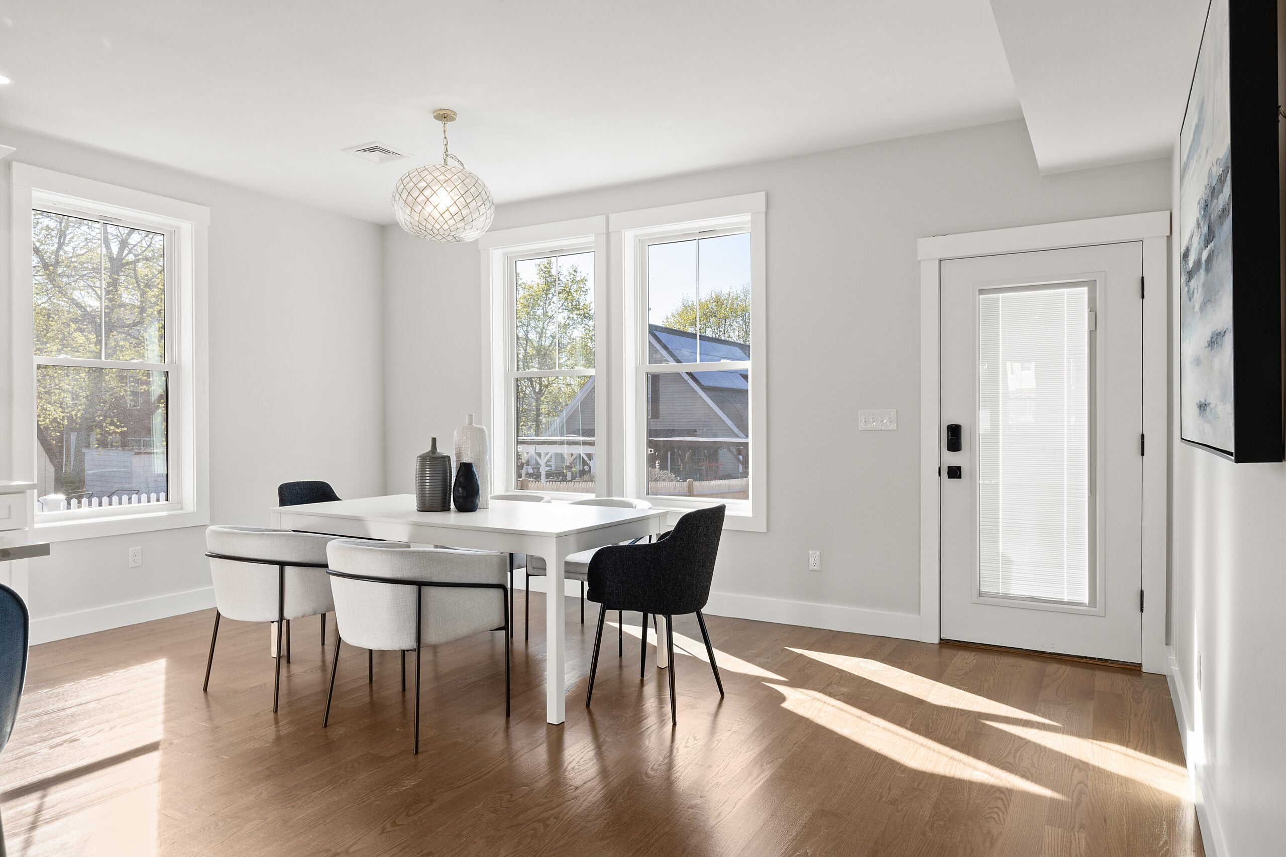 A room with gray walls, three windows (two are paired), a sparkling and bulbous light fixture, a modern table with three Eames-like chairs, and wood flooring. A glass door sits to the back right. Home of the Week in Mattapan.