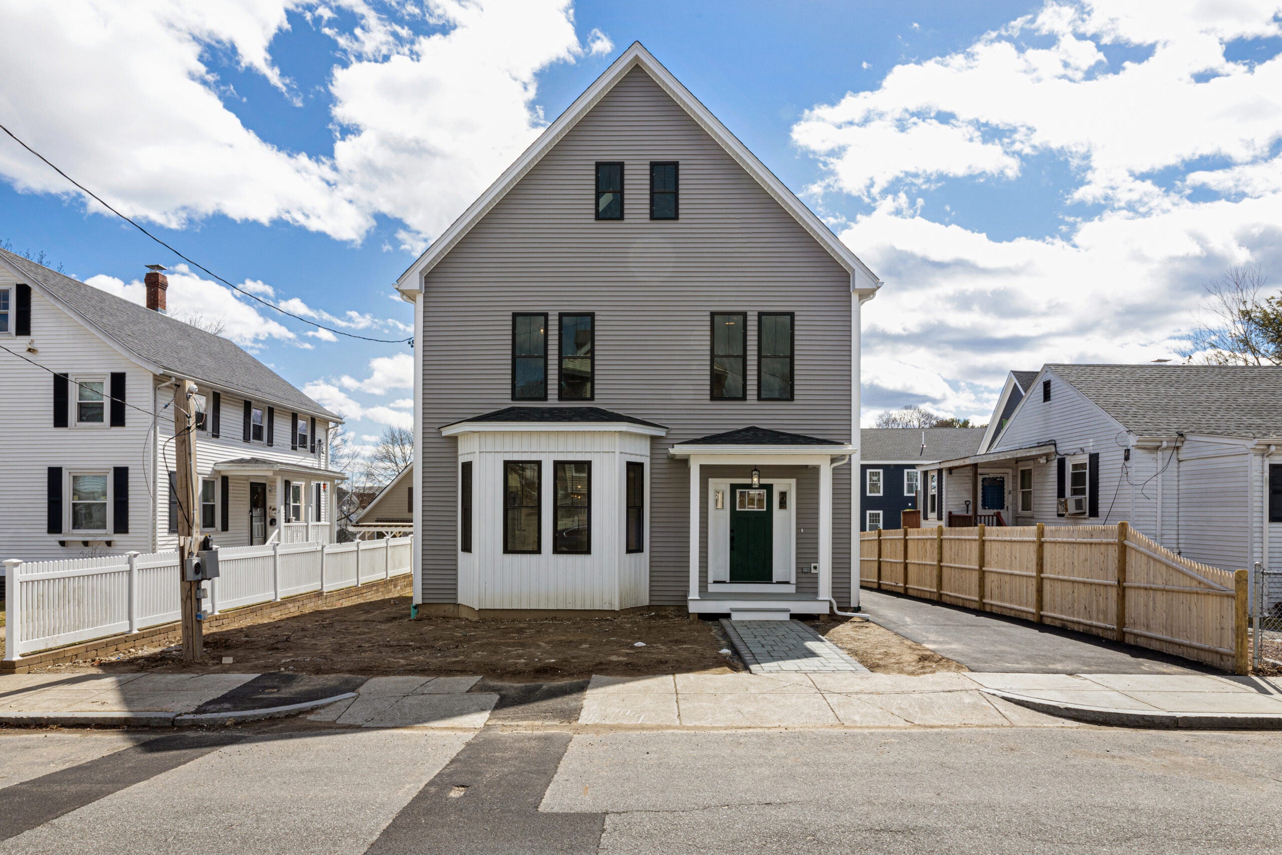 A front-on view of a gray three-story home with an A-frame roof, gray siding, a white bow window bump-out, and a small front porch with a gable roof. A driveway runs along the right side. Fences define the yard. The sky is blue with clouds. Home of the Week in Mattapan.