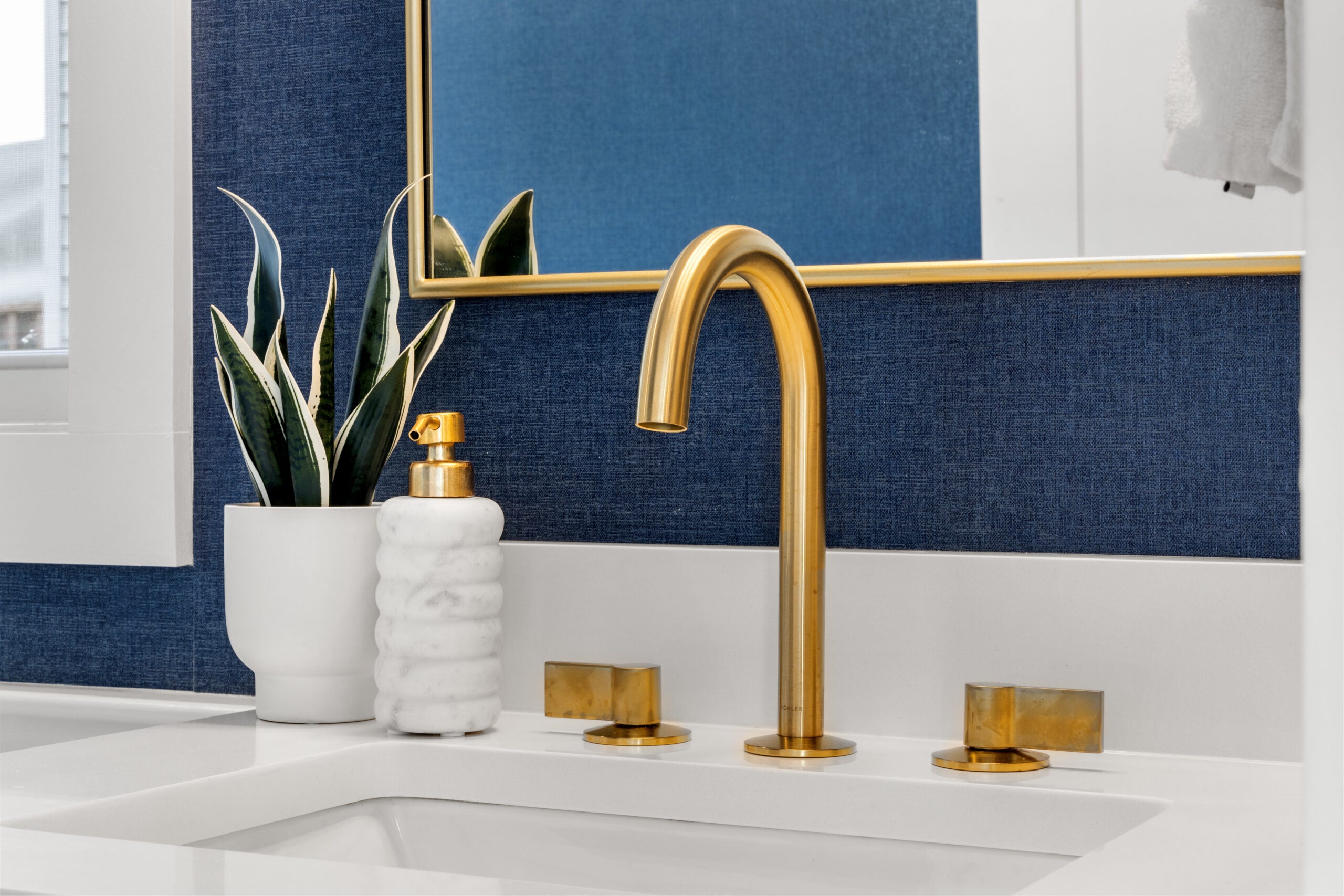 A detail shot of a inset sink with gold-toned fixtures. The faucet itself is long-necked. The wall is navy blue, as is the frame of the mirror hanging on it. A snake plant sits in a white pot. Home of the Week in Mattapan.