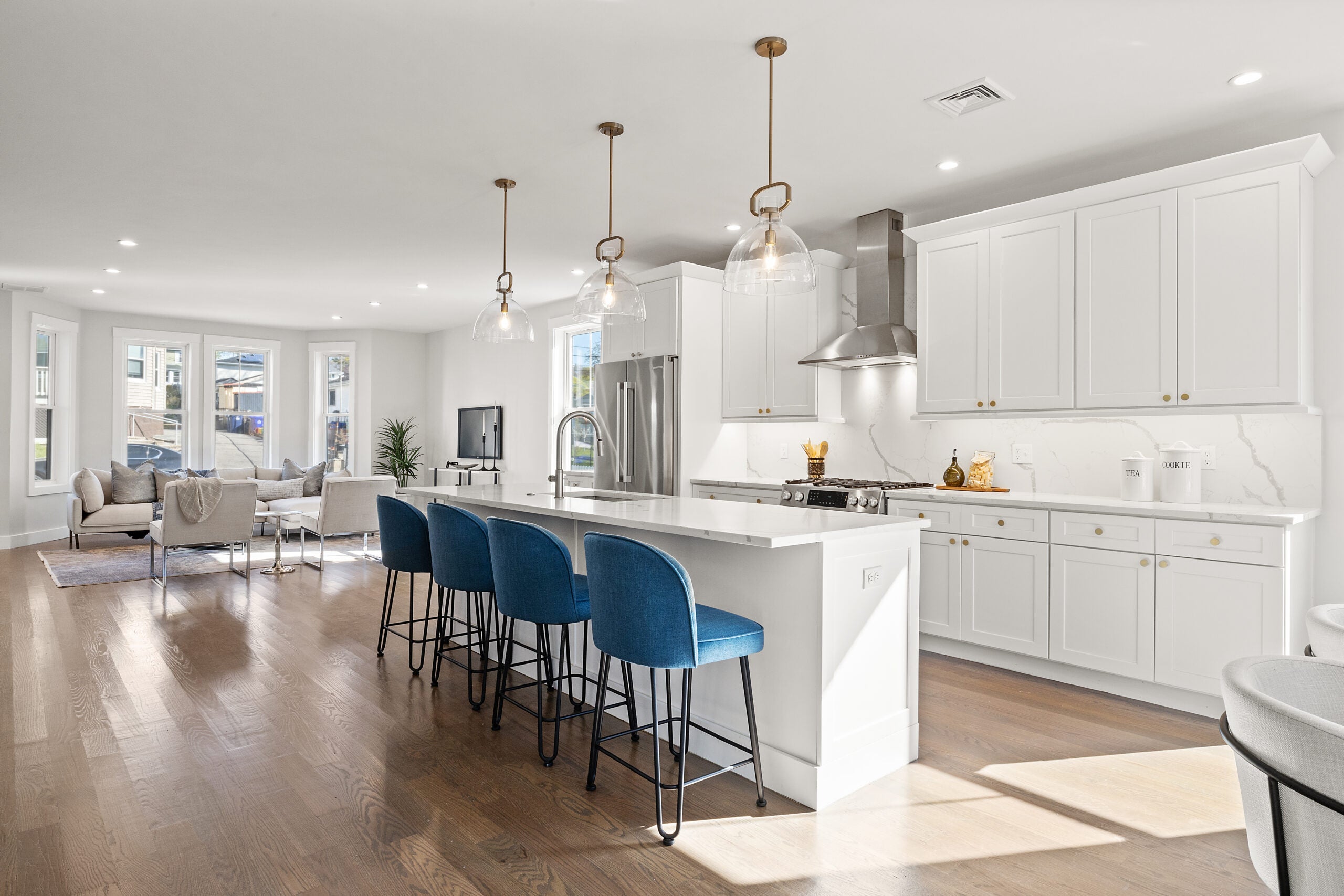 A view from the kitchen into the living area shows a long, white island with a white counter, and blue, backed seating for four. Three globe lights hang above it. The cabinets, which do not extend to the ceiling, are Shaker-style and white, and the appliances are stainless steel. The flooring is a medium-toned wood. Home of the Week in Mattapan.