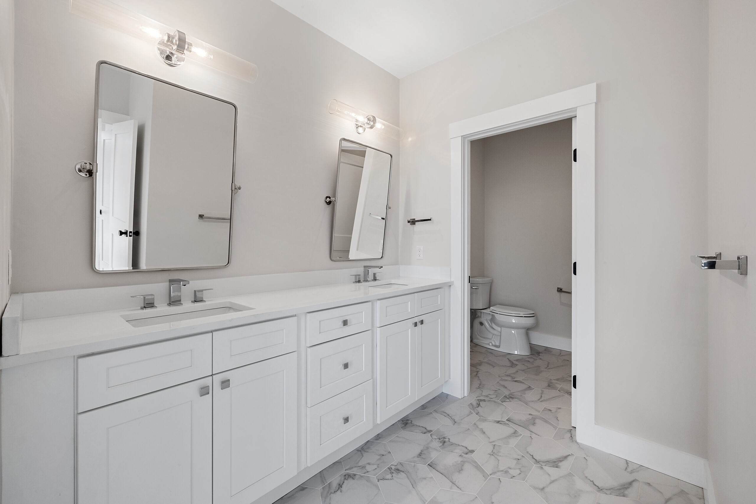 A white double vanity is topped with white quartz. The flooring is a gray and white porcelain tile. The bathroom fixtures are a gray metal. The two square mirrors over the sink tilt toward the user. The water closet is visible in the background. The walls are a soft gray. 