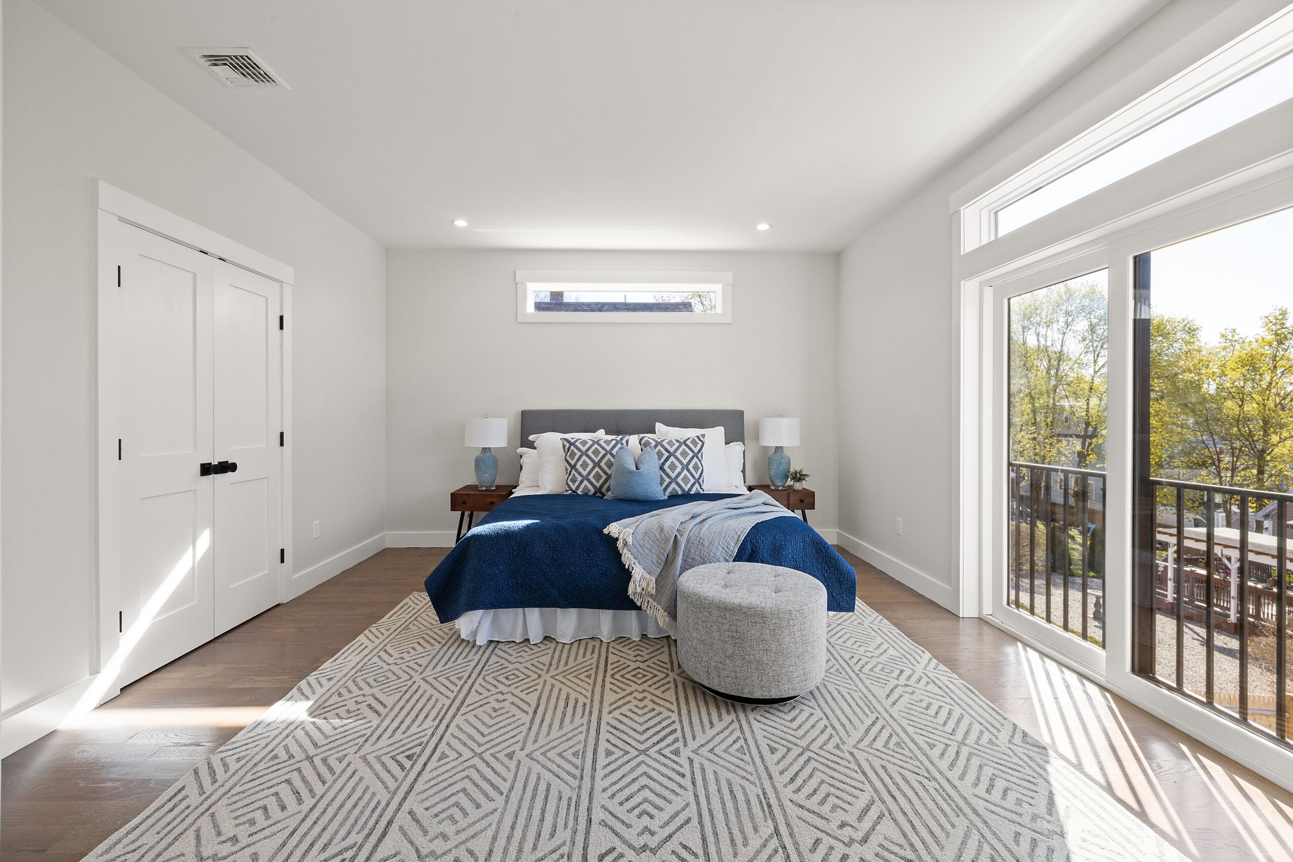 A view of a room with wood flooring, sliders to a Juliet balcony with an iron railing, two casement windows, a gray-and-white patterned area rug, recessed lighting, and soft-gray walls. A king-sized bed with a blue bedspread, a white dust ruffle, and a mix of white and gray pillows sits in the back ground under a casement window. Two night stands with jade-colored lamps flank it. Home of the Week in Mattapan.