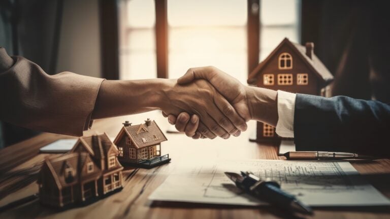 two people of color in suits shaking hands above a desk with blue prints, pens, and model single-family homes.