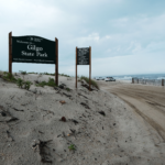 A sign reading "Gilgo State Park" on a sandy beach in view of the ocean.