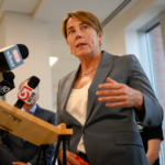 A woman in a grey suit speaks from a podium with multiple microphones.