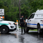 A police officer stands between two vehicles in front of a road that has been barricaded due to flooding.