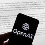 The OpenAI logo is seen on a mobile phone in front of a computer screen.