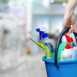 A cleaning woman is standing inside a building holding a blue bucket fulfilled with chemicals and facilities for tidying up in her hand.