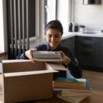 Smiling Indian woman unpacking parcel, holding books, sitting at table, satisfied customer received online store order, delivery service, happy female packing belongings, preparing for relocation, a move.