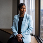 Andrea Cambell sits by a window, smiling and wearing a pale blue blazer.