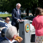 A man in a suit speaks from an outdoor podium to a small crowd